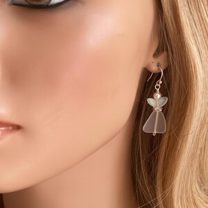 May include: A pair of angel-shaped earrings. The earrings are made of frosted glass with a silver hook. The angel's body is a frosted glass triangle, with a small pearl head and a rhinestone band at the waist.
