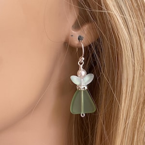 May include: A single earring featuring a small angel design. The angel has a light green dress, a pearl head, and clear wings. The earring is attached to a silver hook. The earring is worn on a person's ear.
