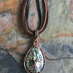May include: A copper wire wrapped pendant featuring a green and blue abalone shell with a tree design. The pendant is attached to a brown leather cord.