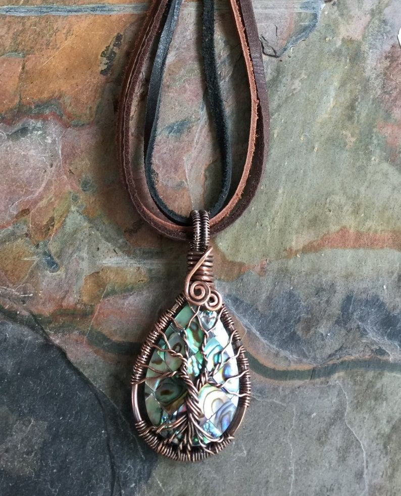 May include: A copper wire wrapped pendant featuring a green and blue abalone shell with a tree design. The pendant is attached to a brown leather cord.