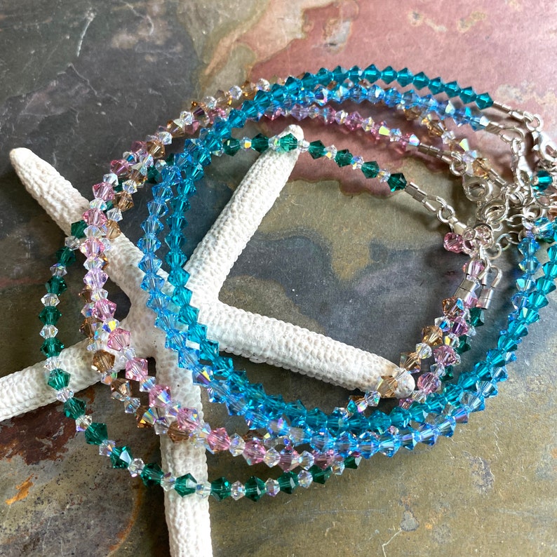 May include: Four delicate crystal bracelets in shades of teal, pink, and green. The bracelets are layered on a white starfish background.
