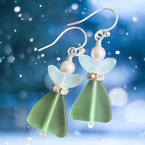 May include: A pair of angel-shaped earrings with silver hooks. Each earring features a light green, triangular body, a light blue wing-like shape, and a pearl bead. The earrings are adorned with small, sparkling rhinestones and are set against a blurred blue background.