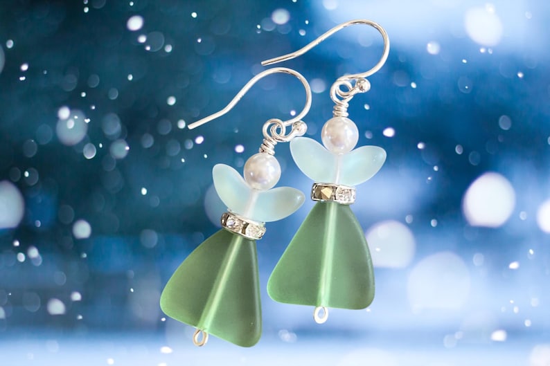 May include: A pair of angel-shaped earrings with silver hooks. Each earring features a light green, triangular body, a light blue wing-like shape, and a pearl bead. The earrings are adorned with small, sparkling rhinestones and are set against a blurred blue background.