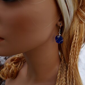 May include: A close-up of a blue glass earring with a silver wire wrap. The earring is a square shape with rounded edges. The earring is hanging from a silver hook. The background is blurred.