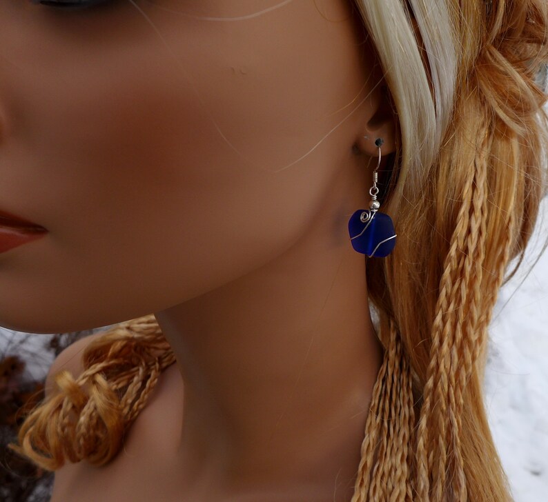 May include: A close-up of a blue glass earring with a silver wire wrap. The earring is a square shape with rounded edges. The earring is hanging from a silver hook. The background is blurred.