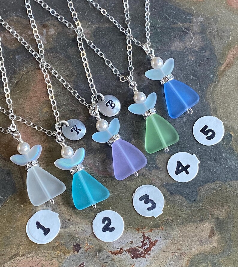 Puede incluir: Cinco collares con forma de &aacute;ngel con cadenas plateadas. Cada collar presenta un colgante de cristal de colores, una perla y un peque&ntilde;o dije plateado. Los colgantes son en tonos azules, verdes y morados. Cada collar tiene un n&uacute;mero en un peque&ntilde;o c&iacute;rculo blanco.