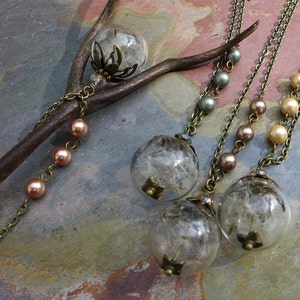 May include: Three necklaces with glass pendants filled with dandelion seeds. The pendants are attached to chains with brown and green beads. The necklaces are displayed on a brown branch.