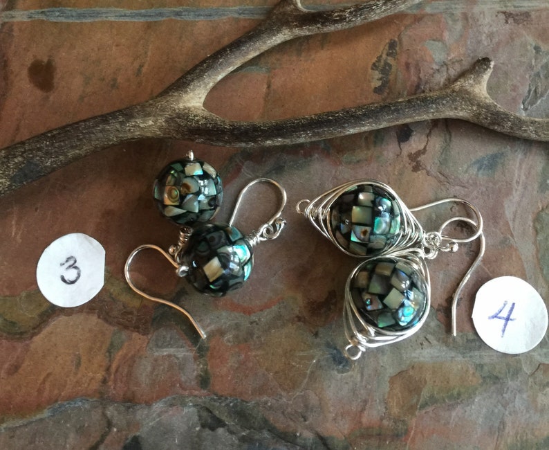 Op de afbeelding: Een paar zilveren oorbellen met zwarte en groene abalone-schelp kralen. De kralen zijn gewikkeld in zilverdraad. De oorbellen bevinden zich op een bruine achtergrond met een witte papieren cirkel met het nummer 3 erop en een andere witte papieren cirkel met het nummer 4 erop.