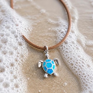 May include: A silver turtle pendant featuring a bright blue iridescent inlay on the shell, suspended from a simple brown leather cord. The turtle charm has a polished metallic finish and is shaped with four flippers and a small head.