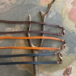 May include: Four leather cords with copper clasps. The cords are brown, orange, black, and brown. The cords are arranged in a row on a wooden surface.