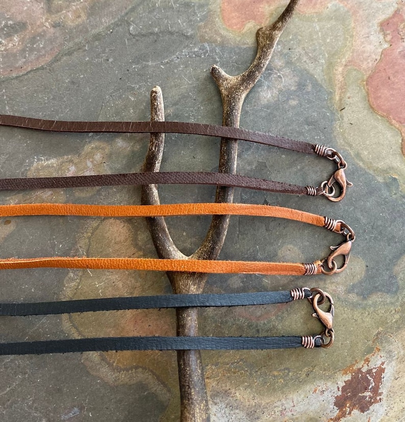 May include: Four leather cords with copper clasps. The cords are brown, orange, black, and brown. The cords are arranged in a row on a wooden surface.