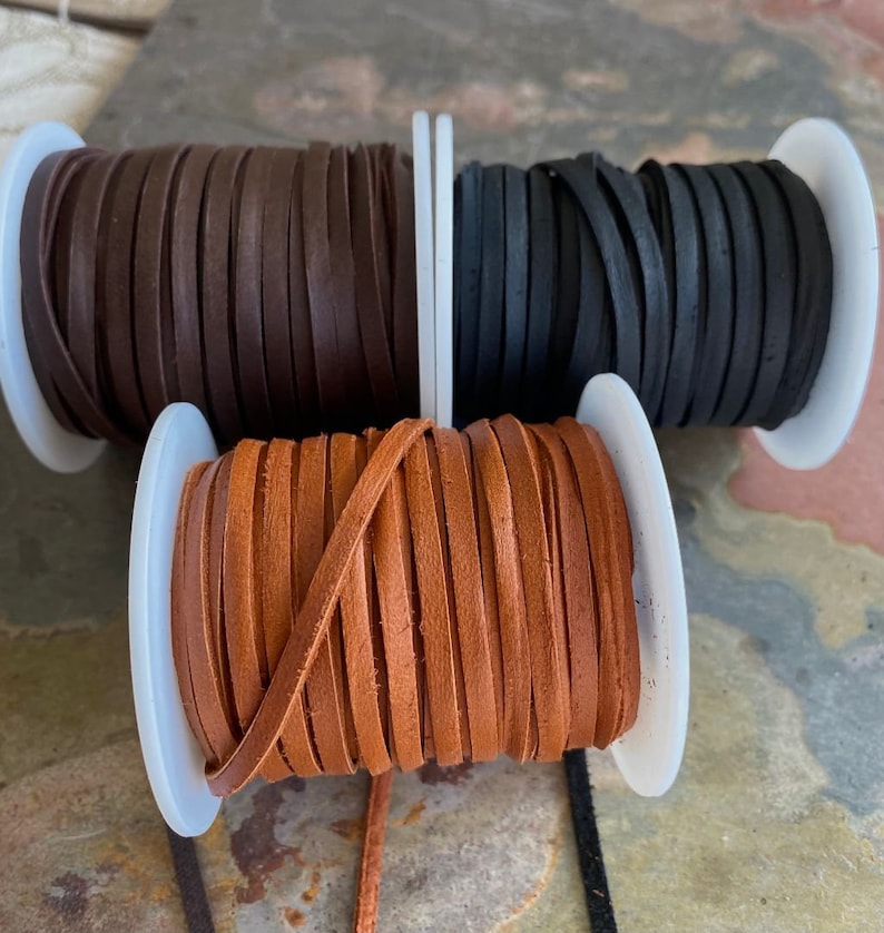 May include: Three spools of leather cord in brown, black, and tan. The cords are wound around white plastic spools.