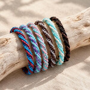 May include: A set of six braided cord bracelets, each featuring a two-tone twisted design. The color combinations include red and blue, purple and light blue, pink and light blue, brown and grey, white and light blue, and solid dark brown.