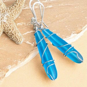 May include: A pair of blue teardrop earrings with silver wire wrapping. The earrings have a hook closure and are displayed on a sandy surface, with a starfish in the background. The earrings are a vibrant turquoise color.