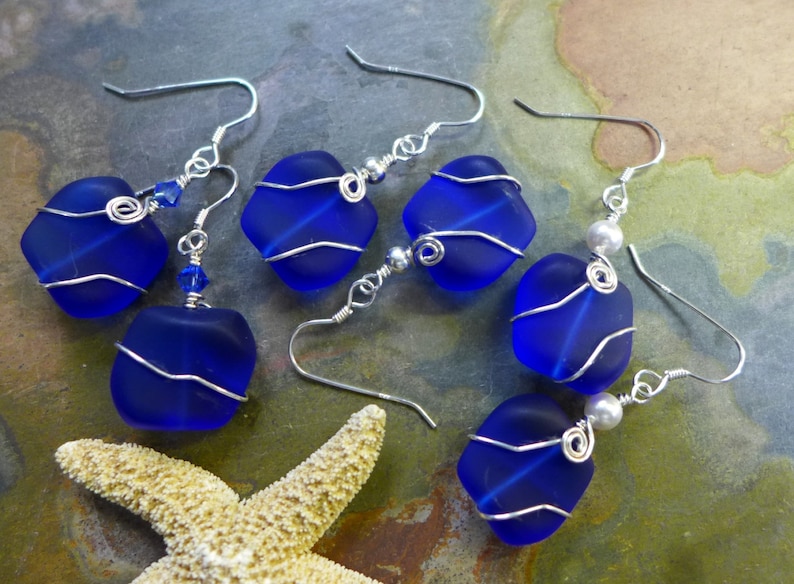 May include: Six pairs of handcrafted earrings featuring vibrant blue sea glass. Each piece is wire-wrapped in silver, with some designs including small blue crystals or pearls. The earrings are displayed with a starfish.