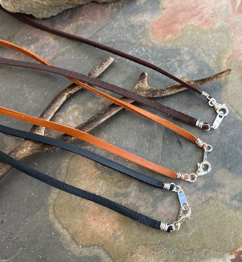 May include: Four leather cords with silver clasps. The cords are brown, orange, black, and dark brown. The cords are arranged in a row, with the brown cord at the top and the dark brown cord at the bottom.