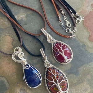 May include: Three wire-wrapped pendants with different colored stones. The stones are a deep blue, a red with white veins, and a red with iridescent streaks. Each pendant is on a leather cord.