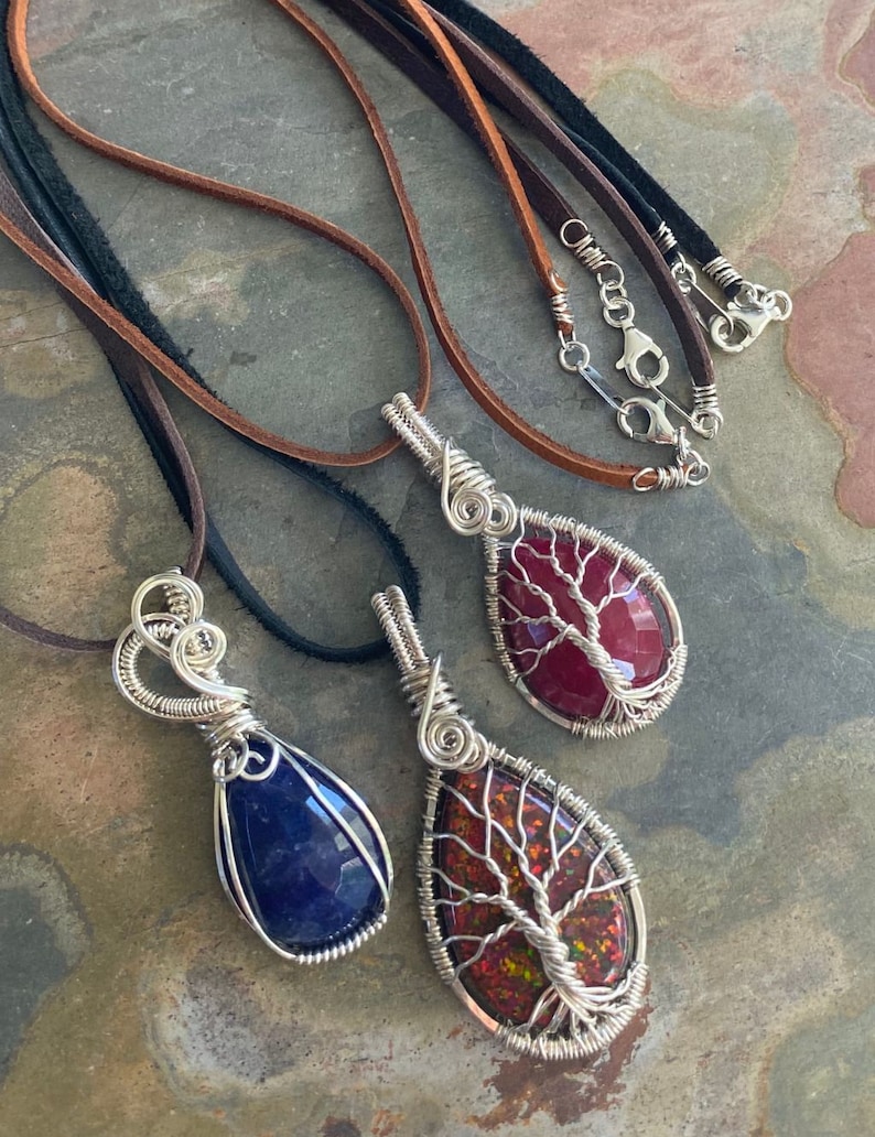 May include: Three wire-wrapped pendants with different colored stones. The stones are a deep blue, a red with white veins, and a red with iridescent streaks. Each pendant is on a leather cord.