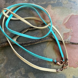 May include: Two leather cords, one turquoise blue and one beige, with copper lobster clasps. The cords are lying on a textured surface.