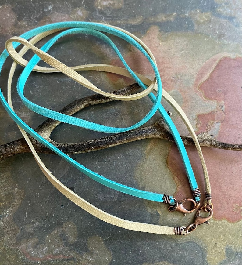 May include: Two leather cords, one turquoise blue and one beige, with copper lobster clasps. The cords are lying on a textured surface.