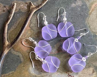 May include: Three pairs of lavender-colored frosted glass earrings, each featuring a square-shaped piece of sea glass wrapped in silver-toned wire. The earrings hang from silver-toned hooks and are accented with small white or purple beads.