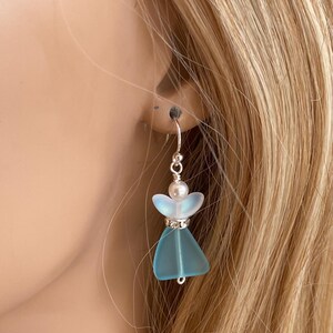 May include: A single earring featuring a small, light blue angel design. The angel has a pearlescent head, a silver-colored halo, and a blue skirt. The earring is suspended from a silver-colored hook.