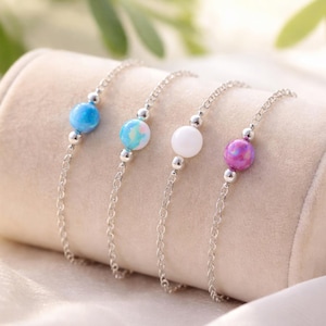 May include: Four delicate bracelets with silver chains and colorful, round beads. The beads are in shades of blue, white, and purple. The bracelets are displayed on a beige, cylindrical stand.