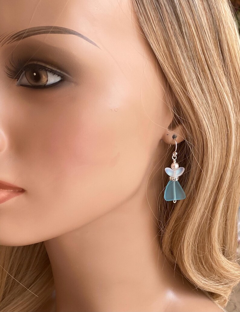 May include: A pair of dangle earrings featuring a light blue angel design. Each earring has a small, pearlescent bead at the top and a silver-colored hook. The angel's wings and dress are a translucent blue color.