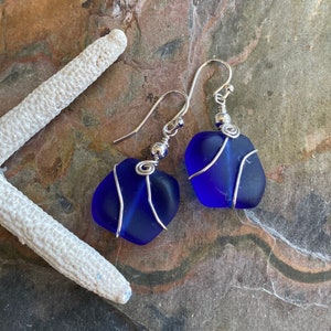May include: A pair of handcrafted earrings featuring vibrant blue sea glass stones. Each stone is wrapped with silver-colored wire and suspended from a silver-tone ear wire. The earrings are displayed on a textured, multi-toned surface, with a white starfish in the background.