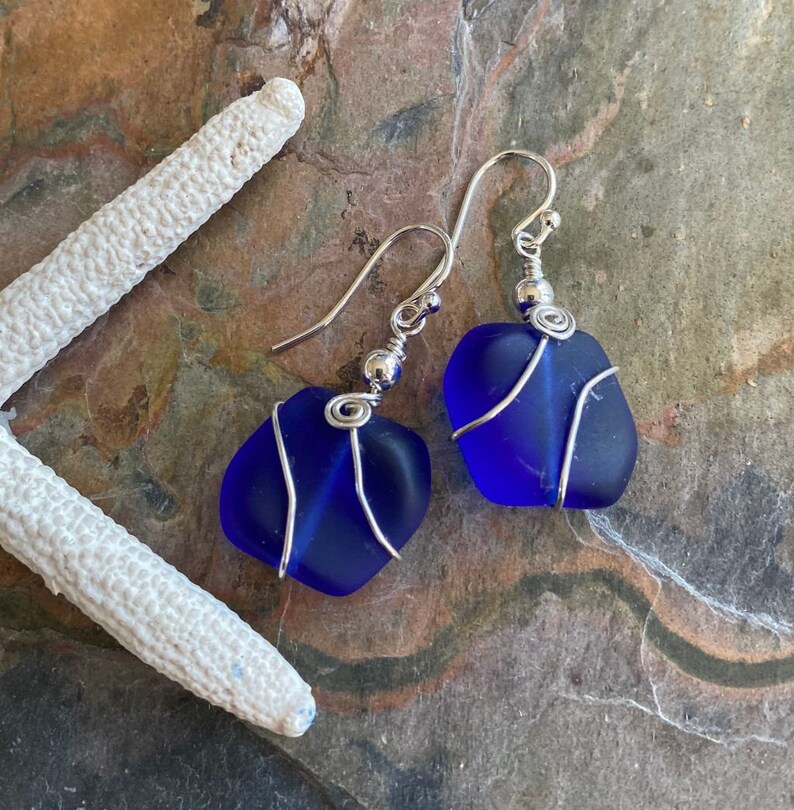 May include: A pair of handcrafted earrings featuring vibrant blue sea glass stones. Each stone is wrapped with silver-colored wire and suspended from a silver-tone ear wire. The earrings are displayed on a textured, multi-toned surface, with a white starfish in the background.