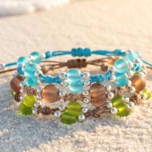 May include: Three handmade bracelets with colorful glass beads and silver accents. The bracelets feature blue, brown, and green beads strung on woven cords. The bracelets are displayed on a sandy surface.