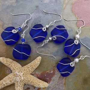 May include: Six pairs of handcrafted earrings featuring cobalt blue sea glass wrapped in silver wire. Some earrings have small blue beads or silver ball accents. A starfish rests near the earrings.