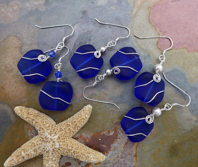May include: Six pairs of handcrafted earrings featuring cobalt blue sea glass wrapped in silver wire. Some earrings have small blue beads or silver ball accents. A starfish rests near the earrings.