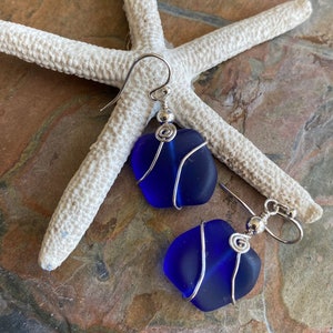 May include: A pair of blue sea glass earrings with silver wire wrapping. The earrings are displayed on a white starfish against a textured, brown and gray surface. The earrings have a simple, elegant design.