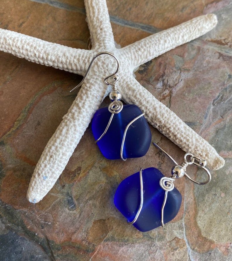 May include: A pair of blue sea glass earrings with silver wire wrapping. The earrings are displayed on a white starfish against a textured, brown and gray surface. The earrings have a simple, elegant design.