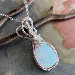 May include: A silver pendant necklace featuring a pear-shaped opal gemstone. The opal is a light, iridescent color with flashes of blue, green, and pink. The gemstone is encased in a wire-wrapped setting and hangs from a delicate silver chain.
