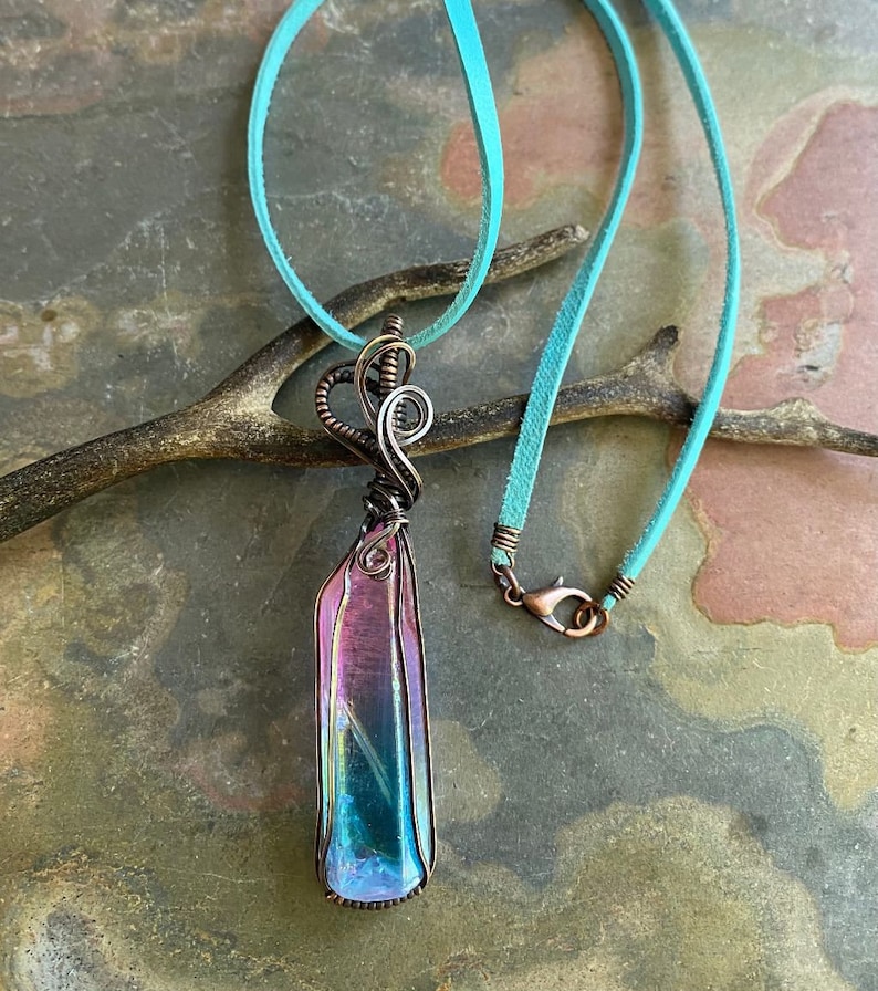 May include: A iridescent crystal pendant wrapped in copper wire, hanging from a turquoise suede cord.