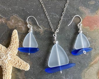 May include: A silver necklace and earrings set featuring sailboat designs. The jewelry includes a pendant and two earrings, each with a translucent sail and a blue base. A starfish is visible in the corner.