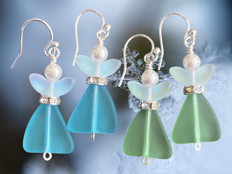 May include: Four pairs of handmade angel earrings. Each earring features a silver hook, a pearl bead, and a decorative crystal band. The angel wings are a light blue or green color, and the body is a matching triangular shape.