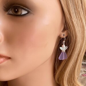 May include: A pair of dangle earrings featuring a small angel design. The earrings have a silver hook and a lavender-colored glass body with a light green head. The angel's dress is a translucent purple color.