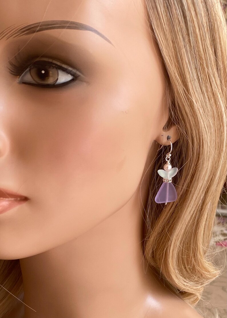 May include: A pair of dangle earrings featuring a small angel design. The earrings have a silver hook and a lavender-colored glass body with a light green head. The angel's dress is a translucent purple color.