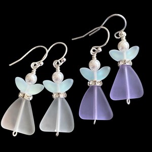 May include: Four pairs of angel-shaped earrings. Each earring features a silver hook, a pearl bead, and a frosted glass body. The wings are light blue, and the dresses are frosted white and lavender. A silver band with rhinestones accents the dress.