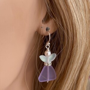 May include: A delicate, handmade earring featuring a lavender-colored glass angel with a silver-colored halo. The angel has a pearl head and a sparkling rhinestone-studded waist. The earring is suspended from a silver-colored hoop.