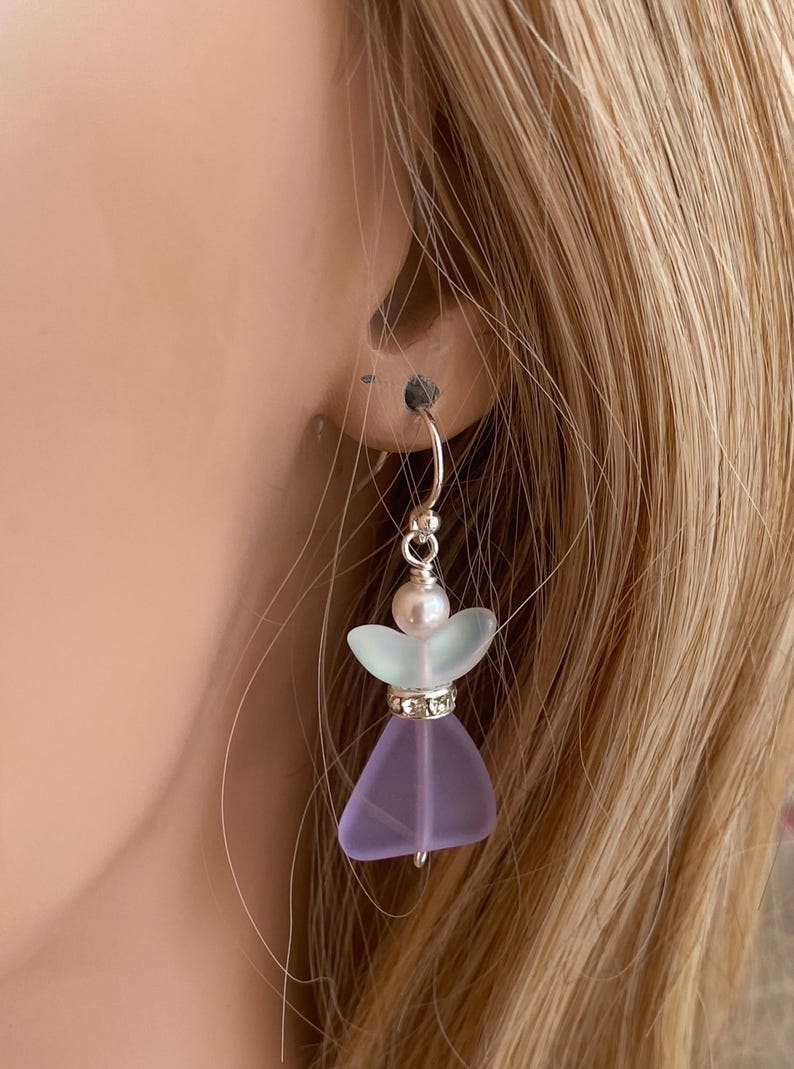 May include: A delicate, handmade earring featuring a lavender-colored glass angel with a silver-colored halo. The angel has a pearl head and a sparkling rhinestone-studded waist. The earring is suspended from a silver-colored hoop.