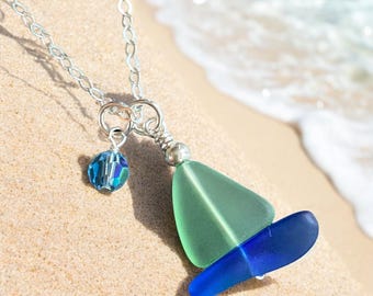May include: A silver chain necklace with a blue and green glass sailboat pendant. The pendant is made of sea glass and has a crystal charm.