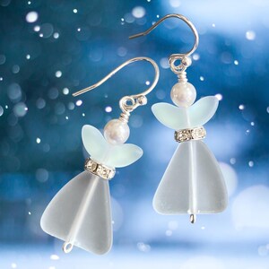 May include: A pair of angel-shaped earrings with silver hooks. Each earring features a frosted glass body, a pearl head, and a rhinestone-studded band. The earrings are set against a blurred blue background with white bokeh.