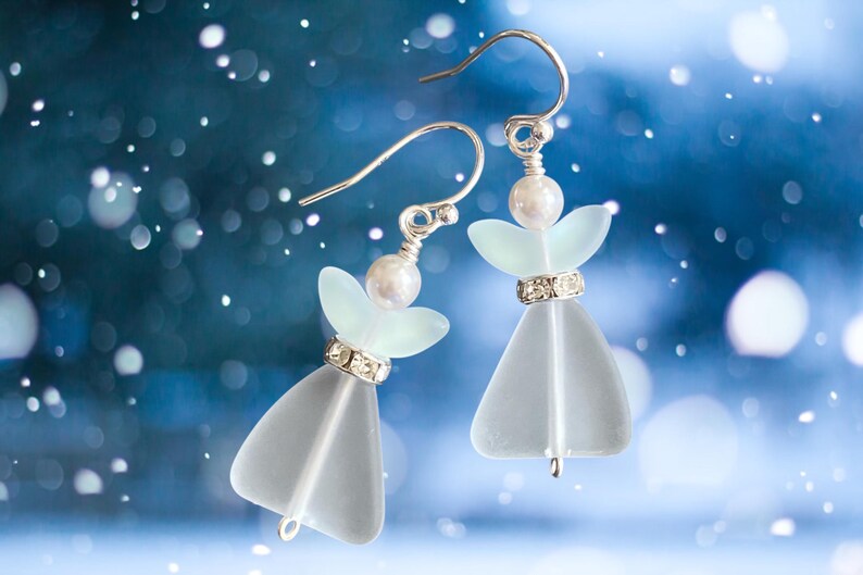 May include: A pair of angel-shaped earrings with silver hooks. Each earring features a frosted glass body, a pearl head, and a rhinestone-studded band. The earrings are set against a blurred blue background with white bokeh.
