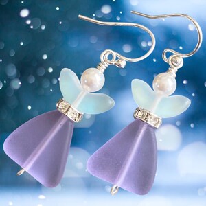 May include: A pair of angel-shaped earrings. Each earring features a lavender-colored, triangular body, a light blue wing design, and a pearl-like bead. The earrings have silver-colored hooks and are adorned with small, sparkling crystals.