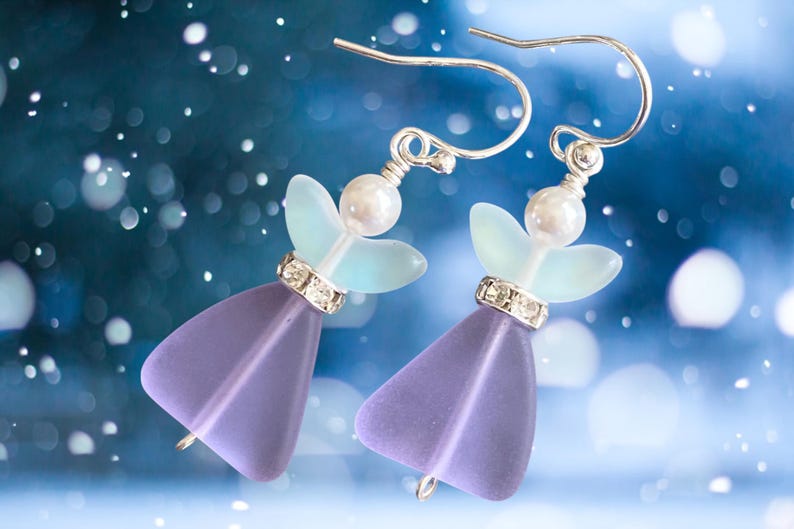 May include: A pair of angel-shaped earrings. Each earring features a lavender-colored, triangular body, a light blue wing design, and a pearl-like bead. The earrings have silver-colored hooks and are adorned with small, sparkling crystals.
