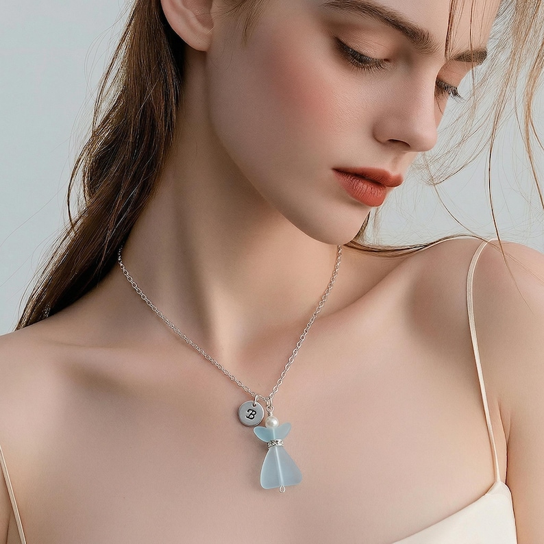 Puede incluir: Un collar plateado con un colgante con un dije en forma de vestido azul claro, una peque&ntilde;a perla y un dije redondo con la letra "B". El collar se lleva alrededor del cuello.